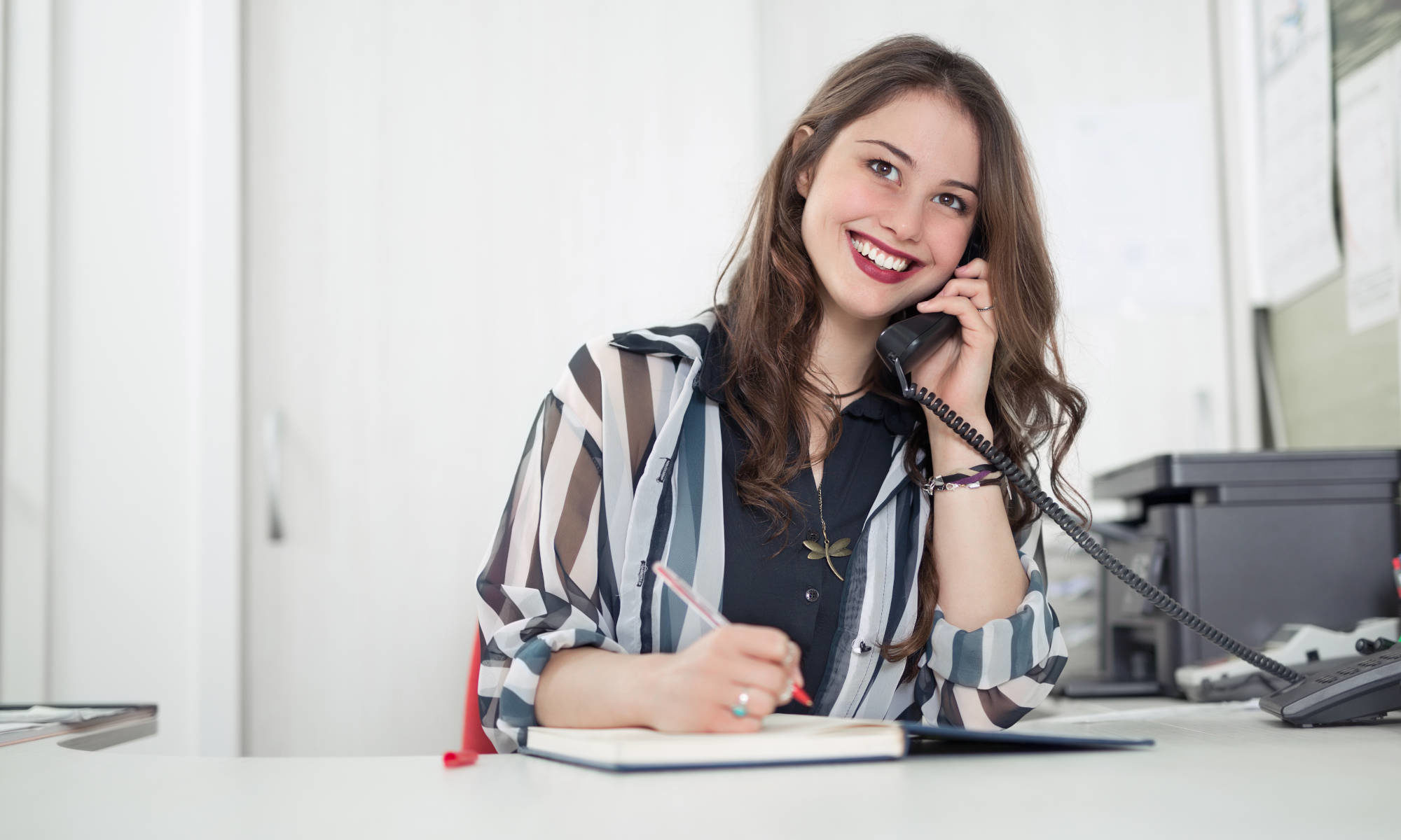 Featured image for "Contact Us" page of Piano Lessons Knoxville dotcom depicting pretty woman in office setting smiling while talking on the office phone and writing in a book.