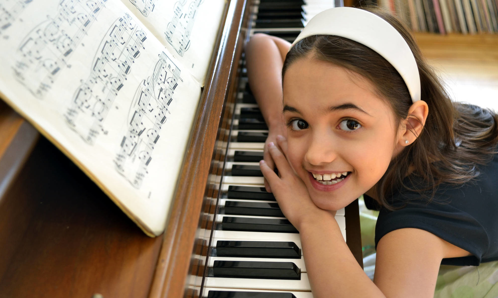 Featured image for "Piano Lessons" page depicting young girl with big smile sitting at piano and looking up at camera.